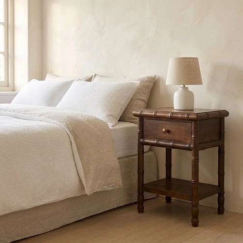 Bamboo Style Bedside Table beside a bed with linen bedding, with a table lamp on top