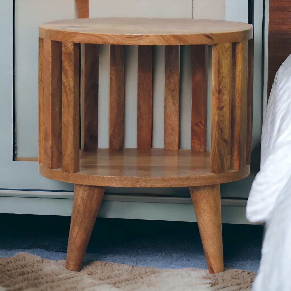 Round bedside table with open shelf, 36 cm wide, light tone, in a bedroom setting next to a bed