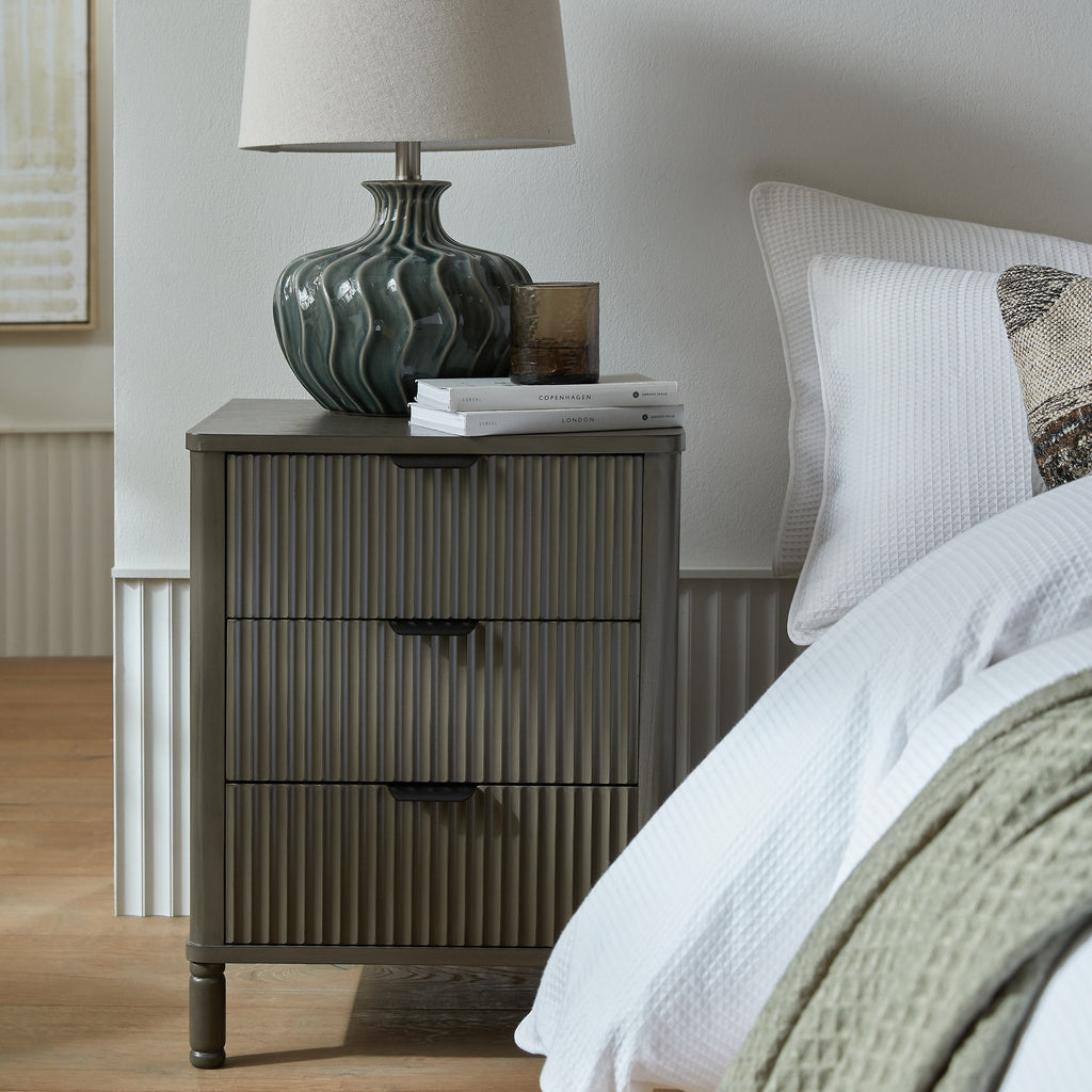 Dark taupe fluted bedside table with 3 drawers beside a bed, with a lamp on top
