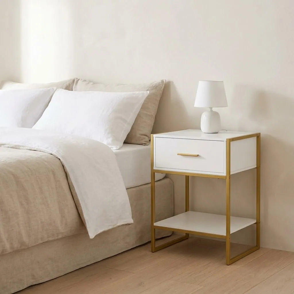 White Bedside Table with Gold Legs beside a bed with linen bedding, with a table lamp on top